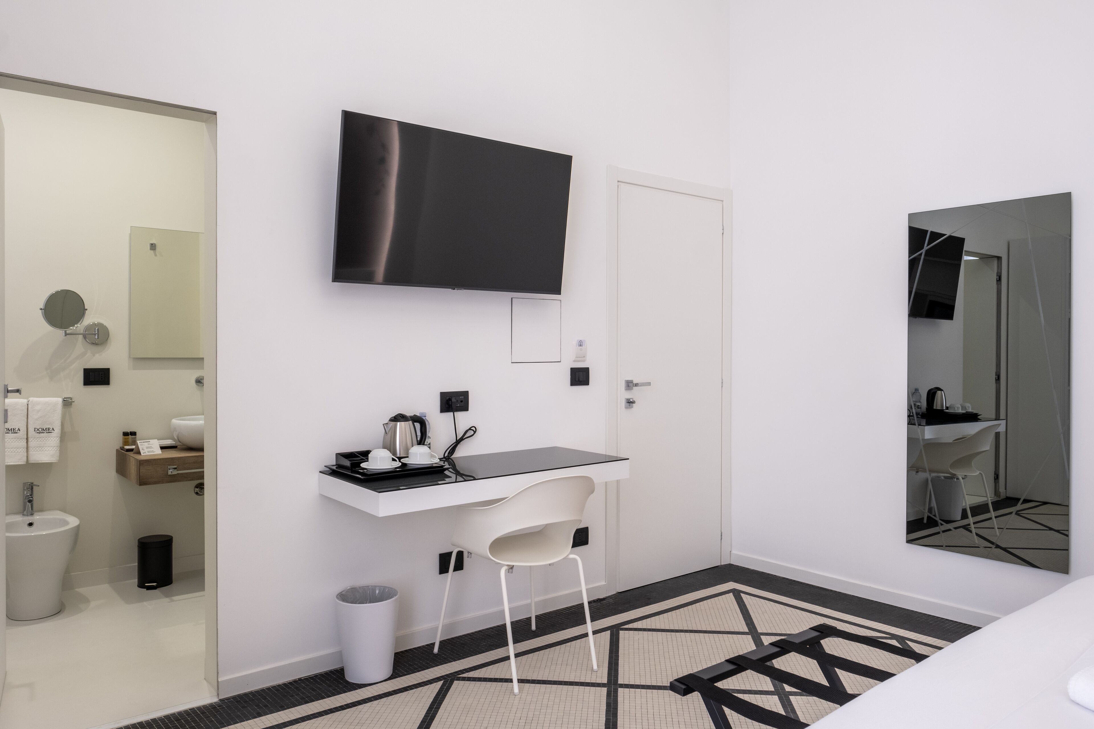 A modern bathroom with a toilet, bidet, and sink. There is a white chair and a small table with a kettle and cups. A flat-screen TV is mounted on the wall, and a mirror is on the opposite wall.