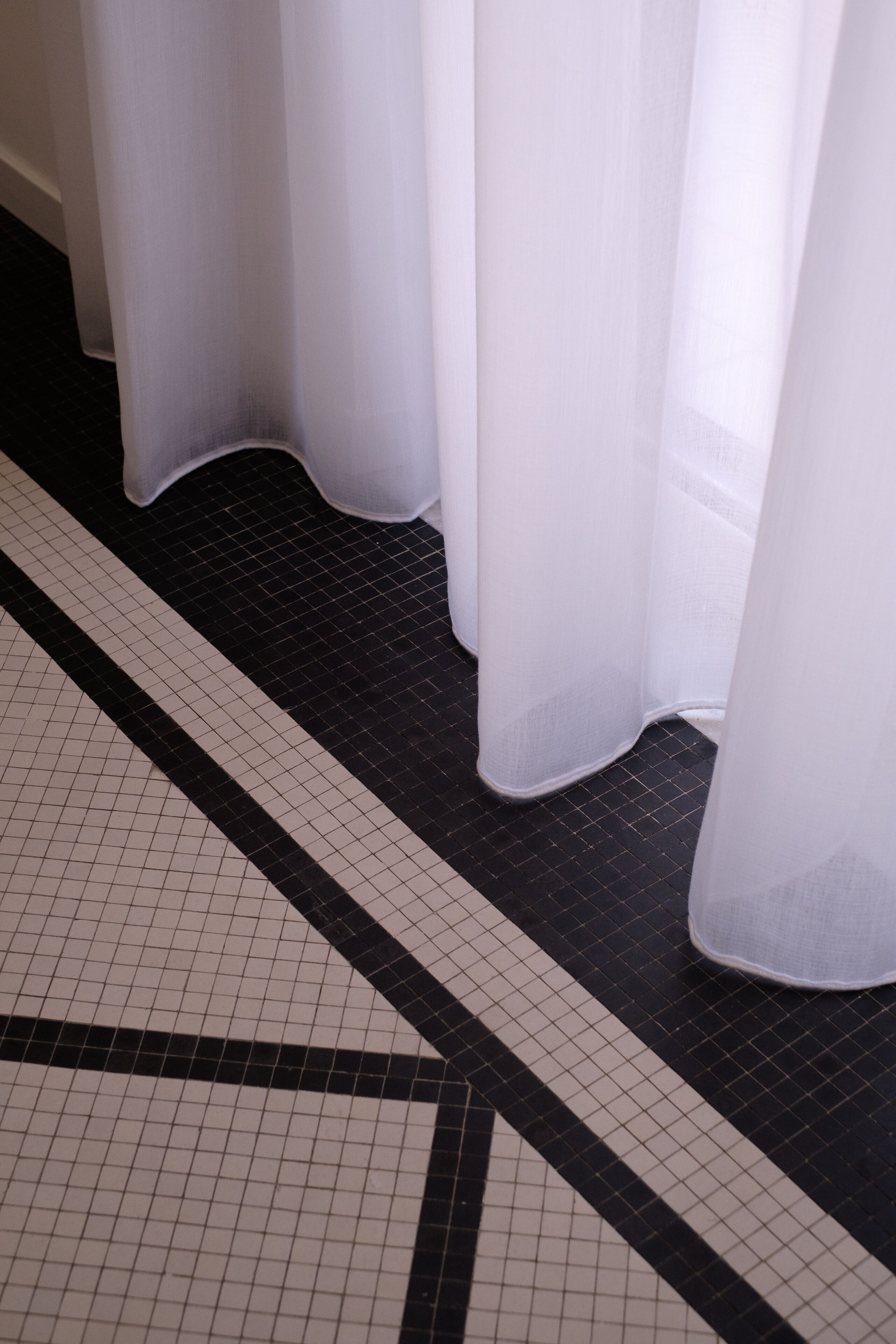 Un sol carrelé orné d’un motif géométrique noir et blanc, des rideaux blancs et une fenêtre partiellement visible.