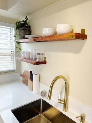 A kitchen with white countertops, a gold faucet, and wooden shelves holding dishes and utensils.