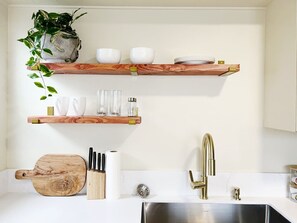 Una cocina con estantes de madera, tabla de cortar, cuchillos, un lavabo y un grifo.