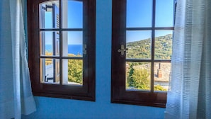 Two windows with wooden frames and white curtains, offering views of a coastal landscape and a hill with buildings. - Apartment Zina Pelion (Zagora-Mouresi)