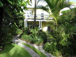 Stroll through the lush garden, surrounded by vibrant greenery and palm trees, to the two-story building with a charming facade.