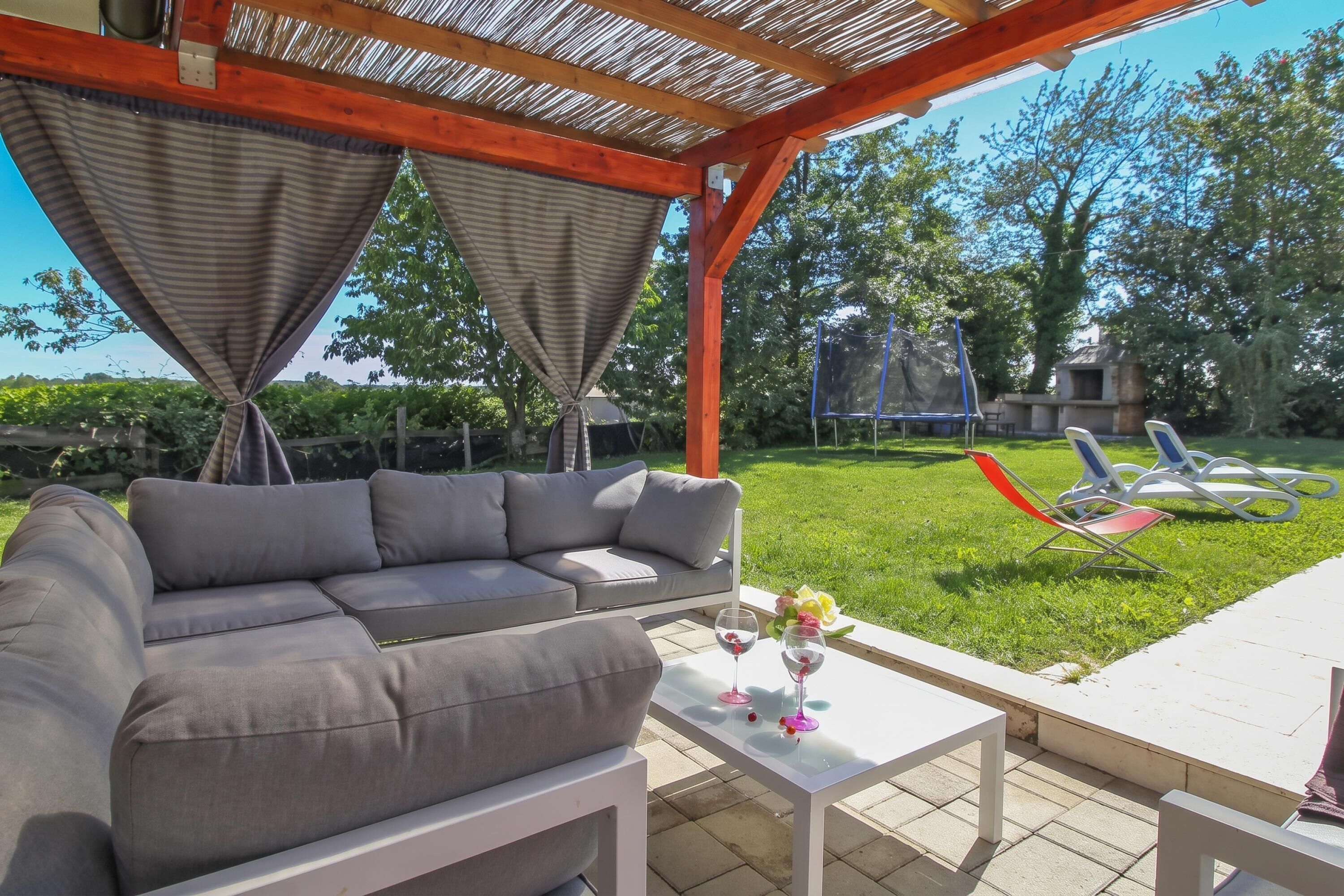 Un espace patio extérieur avec un canapé gris, une table blanche agrémentée de deux verres à vin et d’un vase, et une pergola au-dessus.