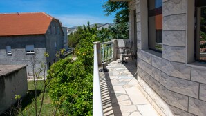 Un balcon doté d’un sol en pierre, d’une table et de chaises, donnant sur un jardin et un bâtiment avec un toit rouge.