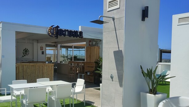 Un espacio de esparcimiento al aire libre con sillas blancas y una terraza de madera, un edificio moderno con entrada de vidrio y un cielo azul despejado.