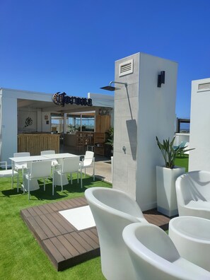 Un espacio de esparcimiento al aire libre con sillas blancas y una terraza de madera, un edificio moderno con entrada de vidrio y un cielo azul despejado.