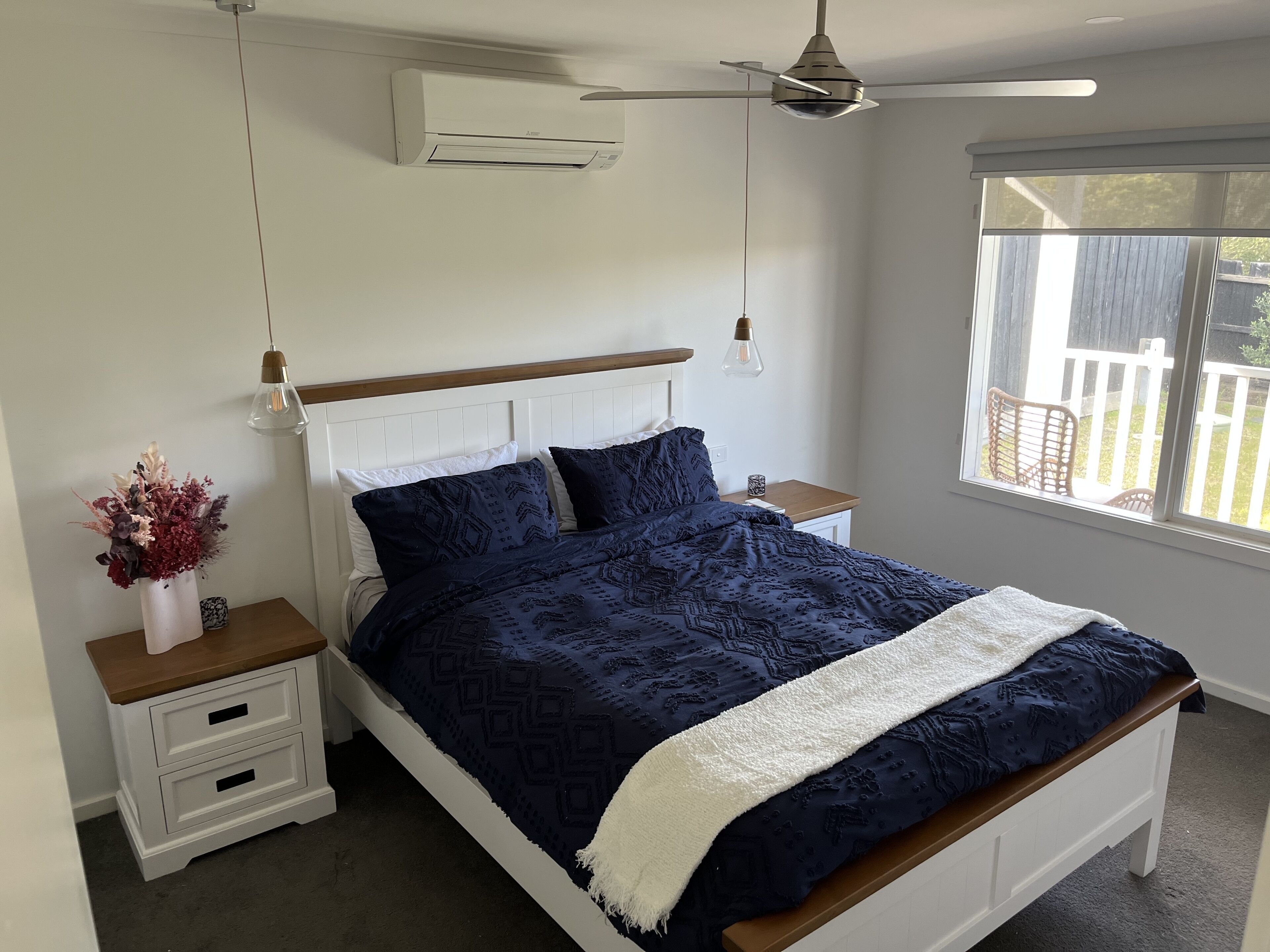 Ein Schlafzimmer mit einem Bett, weißen Nachttischen, einer weißen Kommode, einem Deckenventilator, einer Klimaanlage, einem Fenster mit Blick auf eine Holzterrasse und einer Vase mit Blumen.