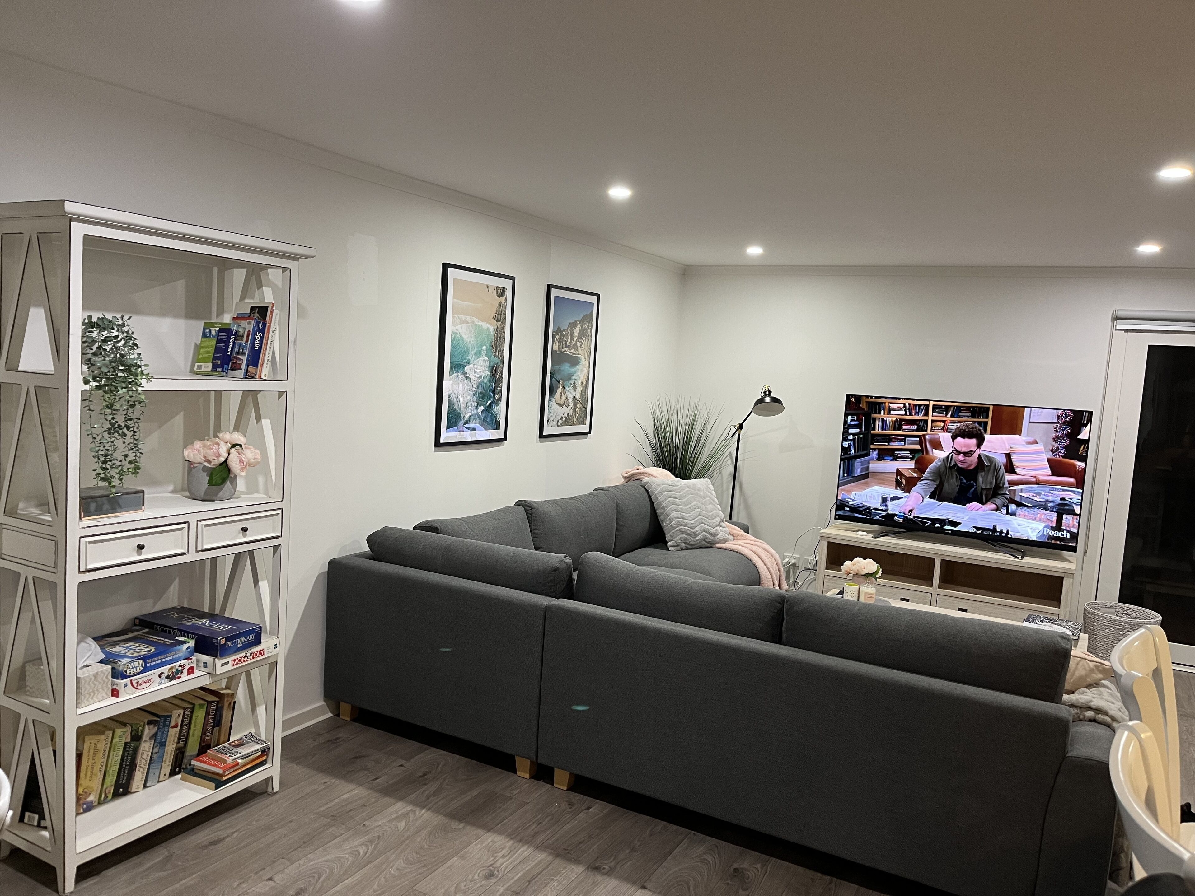 Een moderne woonkamer met een grijze hoekbank, een witte boekenkast met boeken, een flatscreen televisie en ingelijste foto's aan de muur.