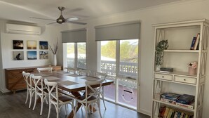 Un coin repas avec une table en bois et des chaises blanches, une bibliothèque blanche contenant des jeux de société et de grandes fenêtres donnant sur une terrasse et des arbres.