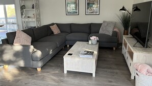 Un salon moderne avec un canapé d’angle gris, une table basse blanche et un téléviseur posé sur un meuble en bois. On peut voir deux tableaux encadrés au mur, une plante et une lampe.