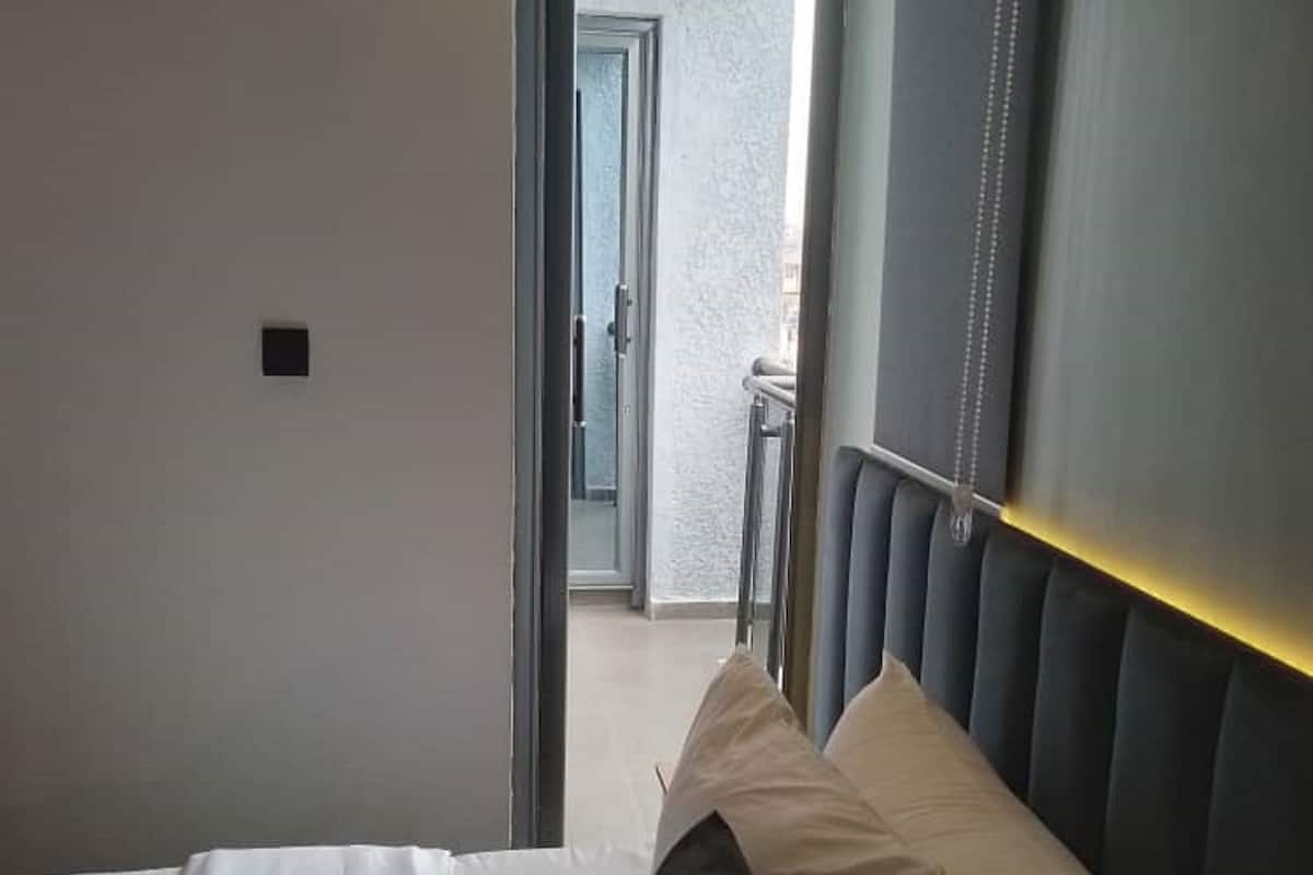 A hotel room with a bed, a headboard with a light strip, and a view of a balcony through an open door.