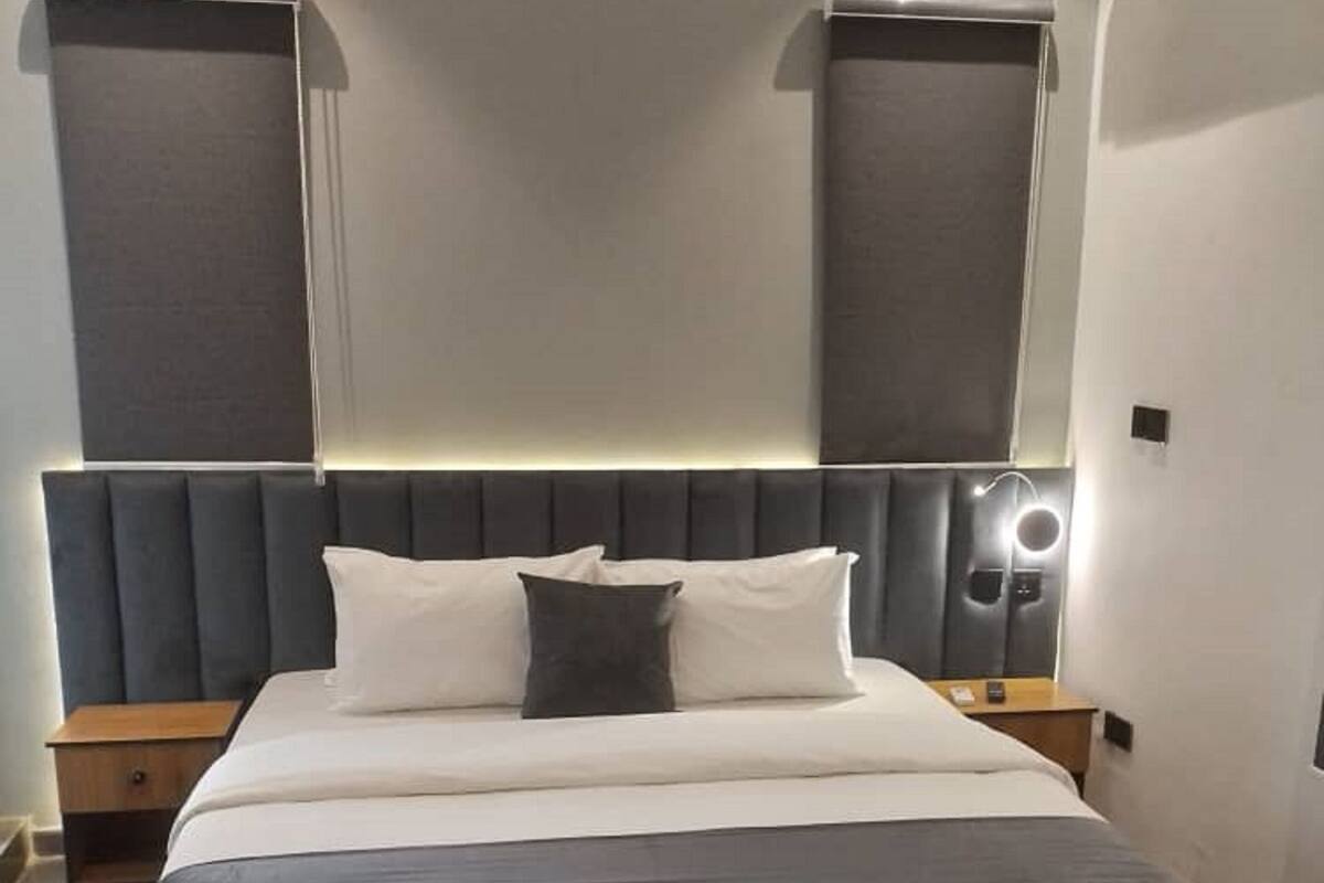 A modern hotel room with a large bed, two bedside tables, a headboard with built-in lighting, and an air conditioning unit on the wall.