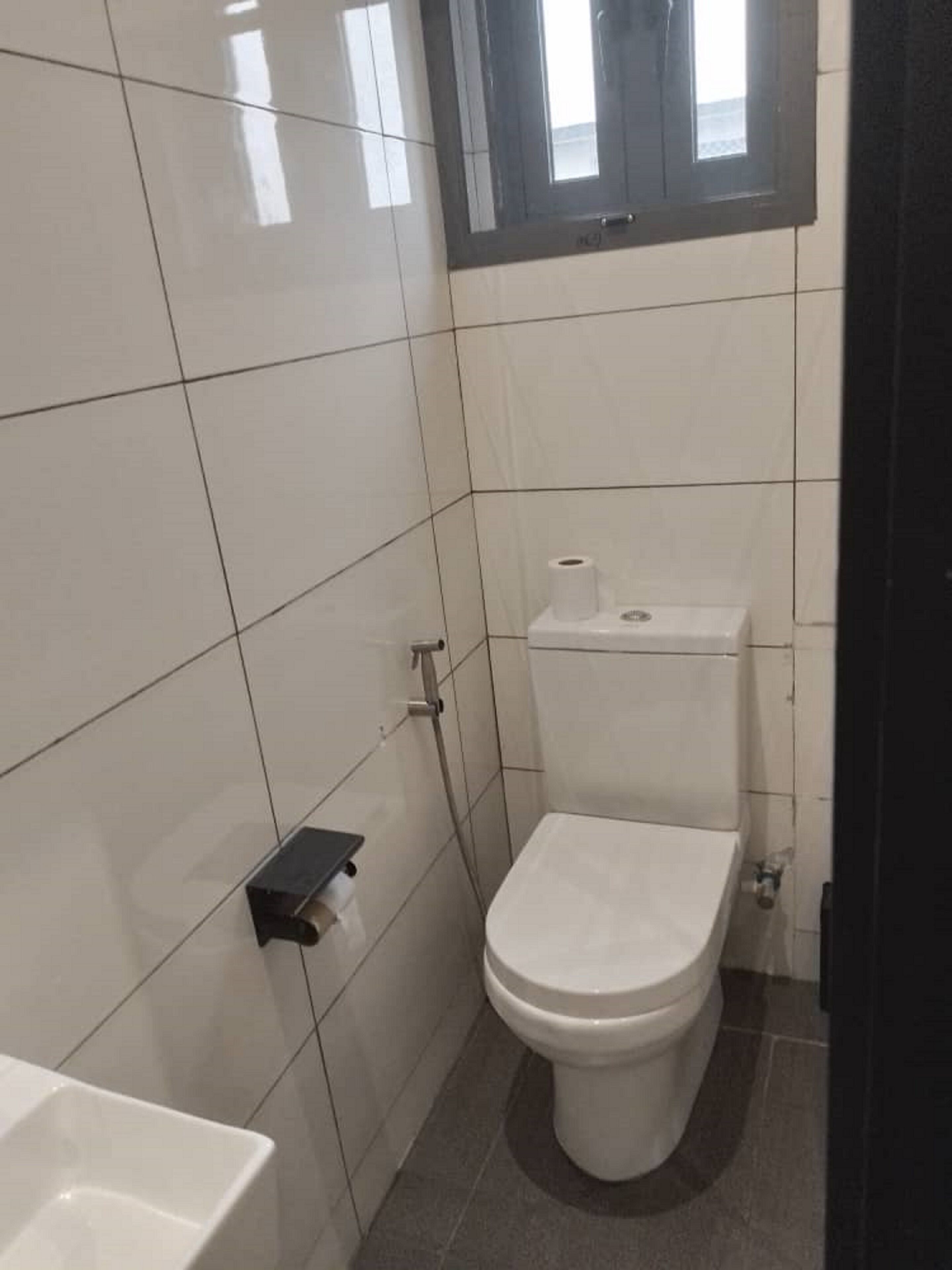 A small, clean bathroom with white tiles, a toilet, and a small shelf with a roll of toilet paper.