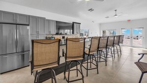 Une cuisine moderne avec des meubles gris, un réfrigérateur en acier inoxydable, un îlot central avec des chaises et un ventilateur de plafond.