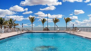 Une piscine rectangulaire à l’eau d’un bleu cristallin, entourée de palmiers et de chaises longues, offrant une vue sur l’océan et un ciel dégagé.