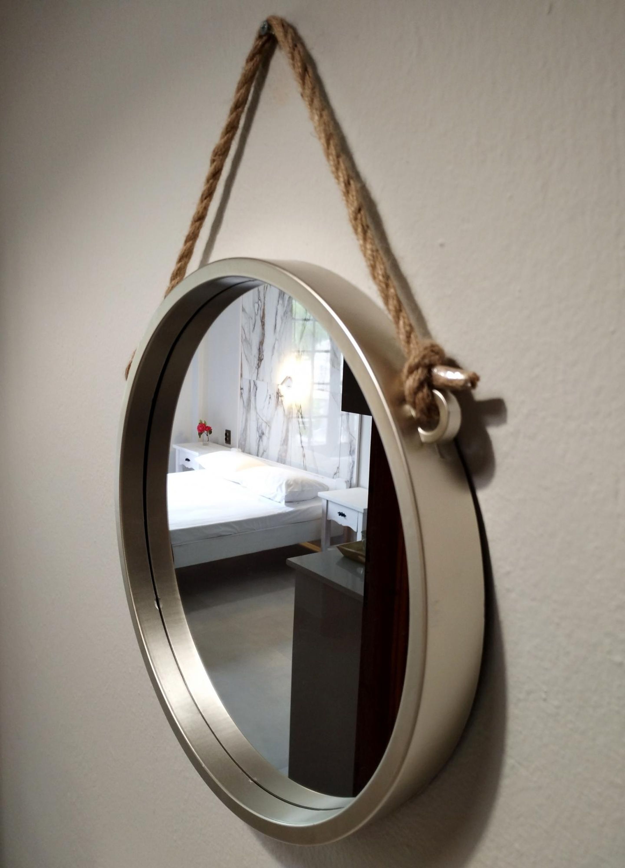 A round mirror with a rope handle, reflecting a bedroom with a bed, bedside table, and a nightstand.