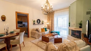A living room with a sofa, coffee table, and a fireplace. There is a dining area with a table and chairs. The room has a chandelier and a view of the city through large windows. - Armonia Rentals Volos Flat (Volos)
