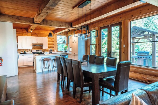 A wooden cabin interior with a dining area, kitchen, and large windows.