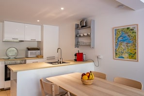 Et moderne køkken med hvide skabe, en træbordplade og et spiseområde med bord og stole. Der er en skål med frugt på bordet og et kort på væggen.