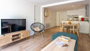 A modern living room with a flat-screen TV, a wooden entertainment unit, a black wire chair, a teal sofa, and a wooden coffee table with a magazine and remote control.