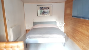 A bedroom with a bed, a bedside table with a lamp, a radiator, and a framed picture on the wall. - Maisonnette (Chambéry)
