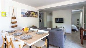 A modern living room with a dining area, grey sofa, flat-screen TV, and a view of the sea through the window.