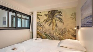 A hotel room with a bed, a window with curtains, a tropical mural on the wall, and a painting of a sunset.