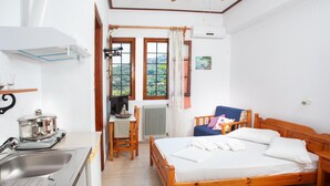A compact room with a bed, a small table with a chair, a kitchen area with a sink, and a window with a view of greenery. - Paradeisos Double Room With Sea View 4 (Zagora-Mouresi)