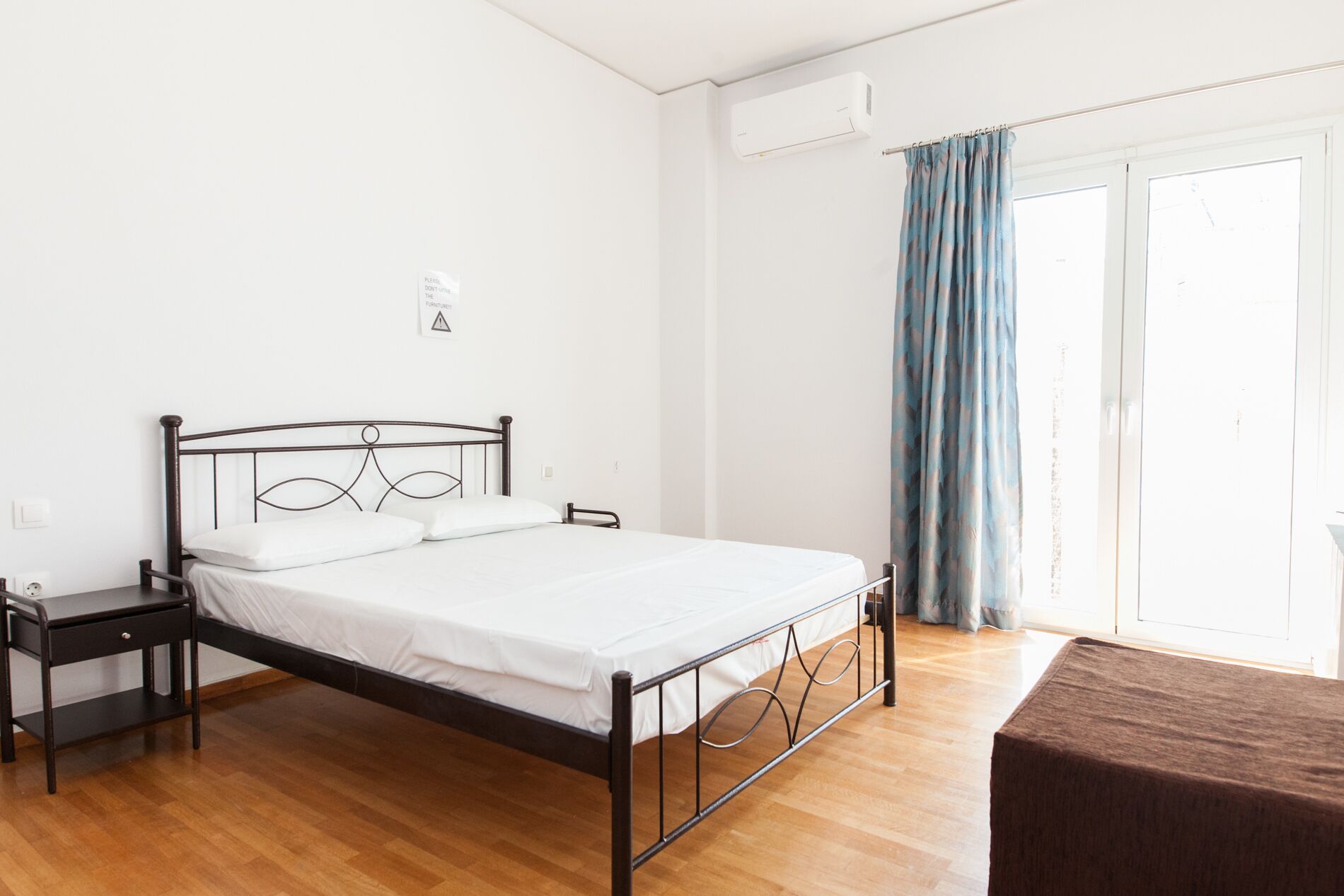 Une chambre avec un lit en métal, une table de chevet, une fenêtre avec des rideaux bleus et un appareil de climatisation fixé au mur.