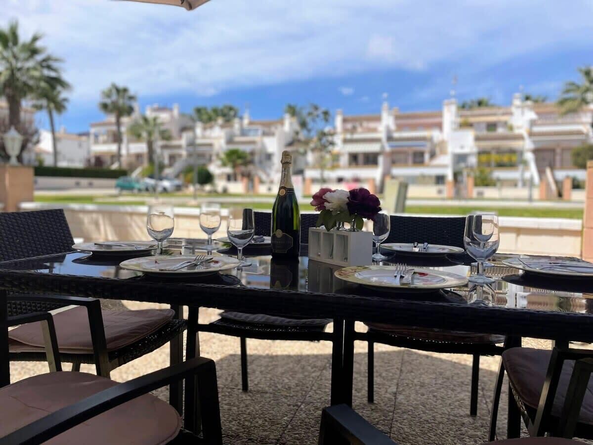 An outdoor dining area with tables set for two, a bottle of wine, and a vase with flowers.