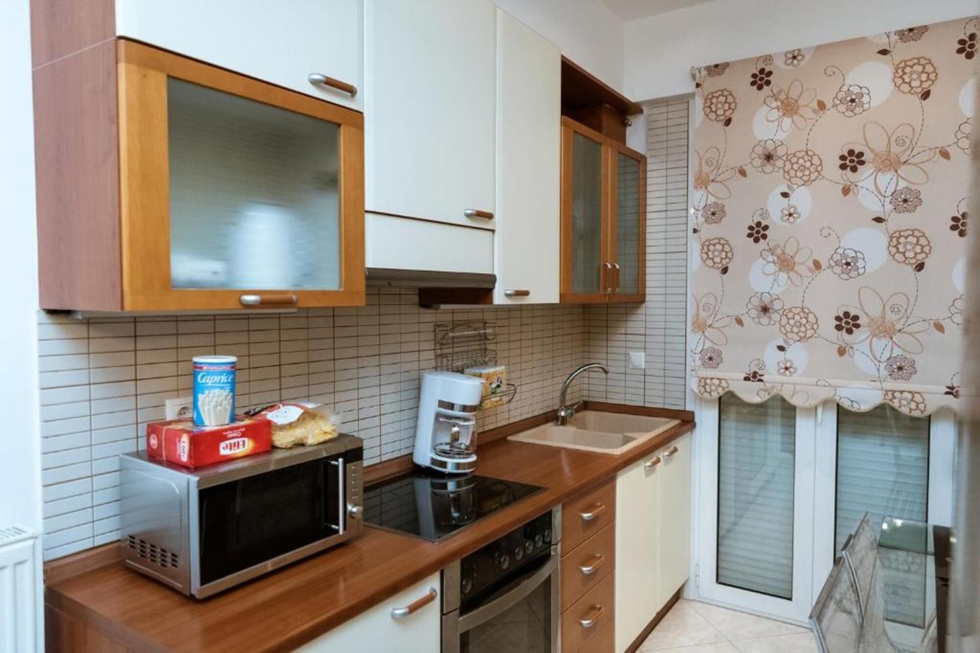 A compact kitchen with a microwave, toaster, and coffee maker on the counter, wooden cabinets, and a window with floral curtains.