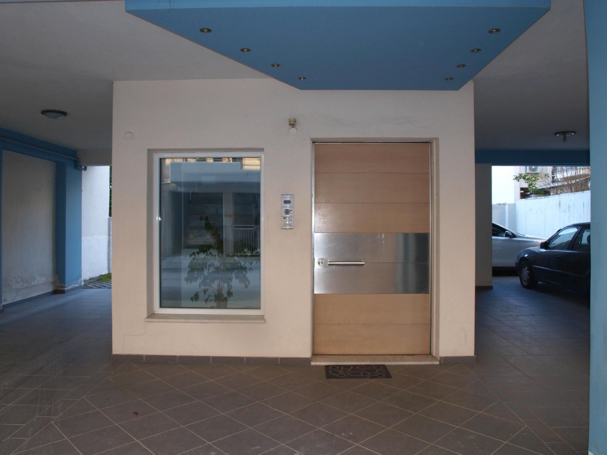 A modern building entrance with a glass door and a wooden door, a tiled floor, and a blue ceiling.