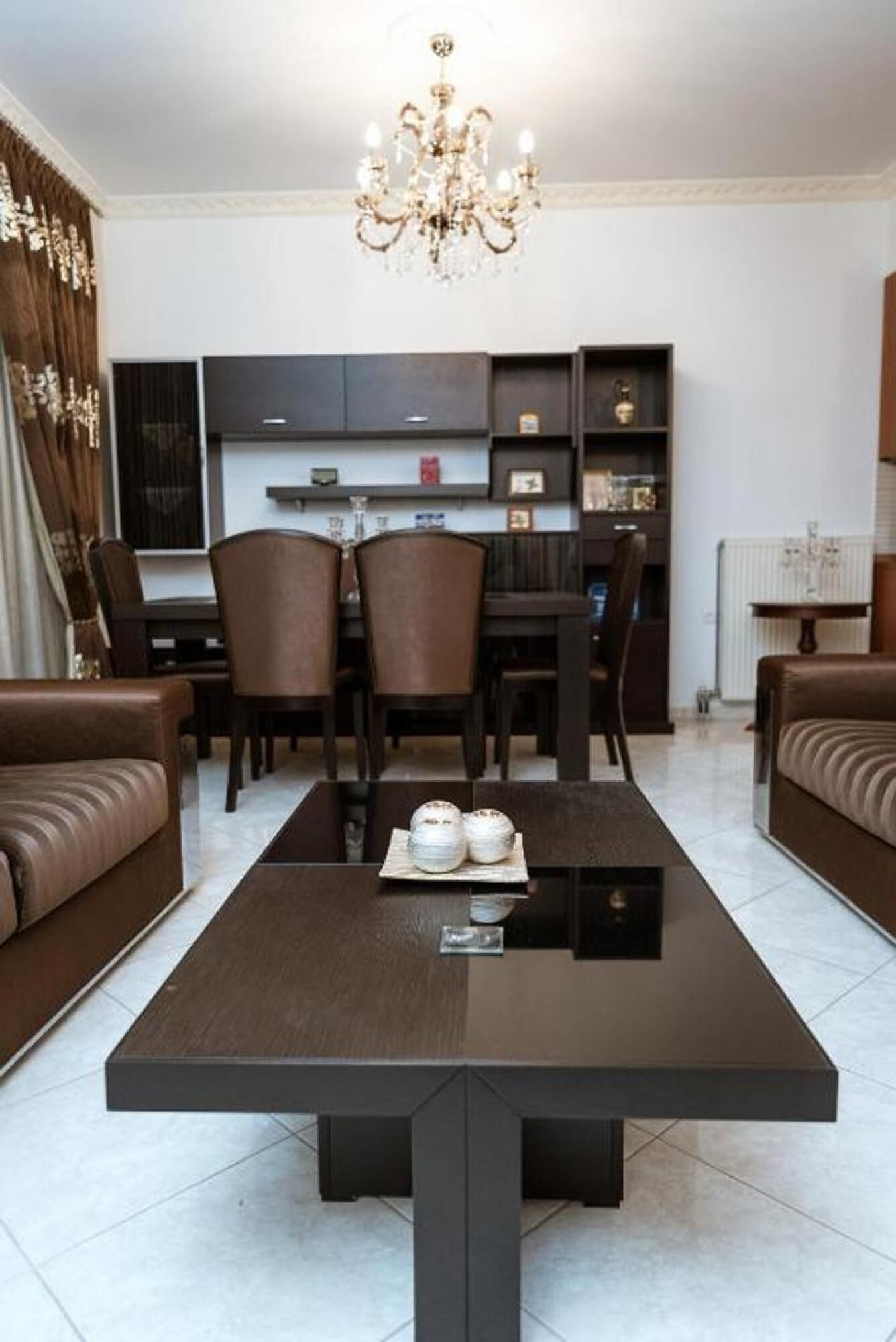 Ein modernes Wohnzimmer mit einem dunklen Holztisch, zwei braunen Ledersofas, einer Kristallleuchtre und einer Küche im Hintergrund.