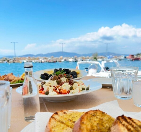 A table with a seafood salad, grilled bread, and a bottle of olive oil, overlooking a marina with boats and a clear sky.