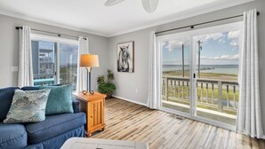 A room with a blue sofa, a wooden side table with a lamp, a ceiling fan, and large windows with a view of the sea and a pier.