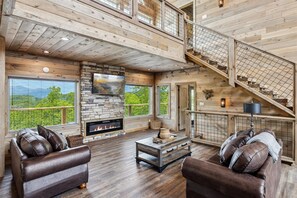 A spacious living room with a fireplace, leather sofas, a coffee table, and a stone accent wall. A staircase with a railing leads to an upper level. - Southern Comfort 5 Bedroom Cabin by RedAwning (Sevierville)