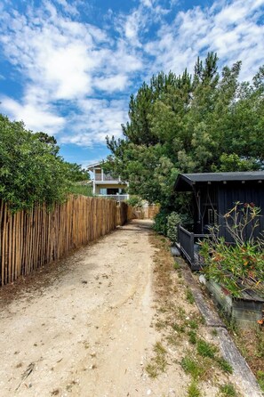 Un sentier de terre mène à un bâtiment à deux étages doté d’un balcon, entouré d’une végétation luxuriante et d’une clôture en bois.