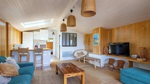 A modern living room with a sofa, coffee table, and TV, wooden walls, and a kitchen in the background. - Villa des Sables: Cosy Apartment with Bassin View and Balcony (Lège-Cap-Ferret)