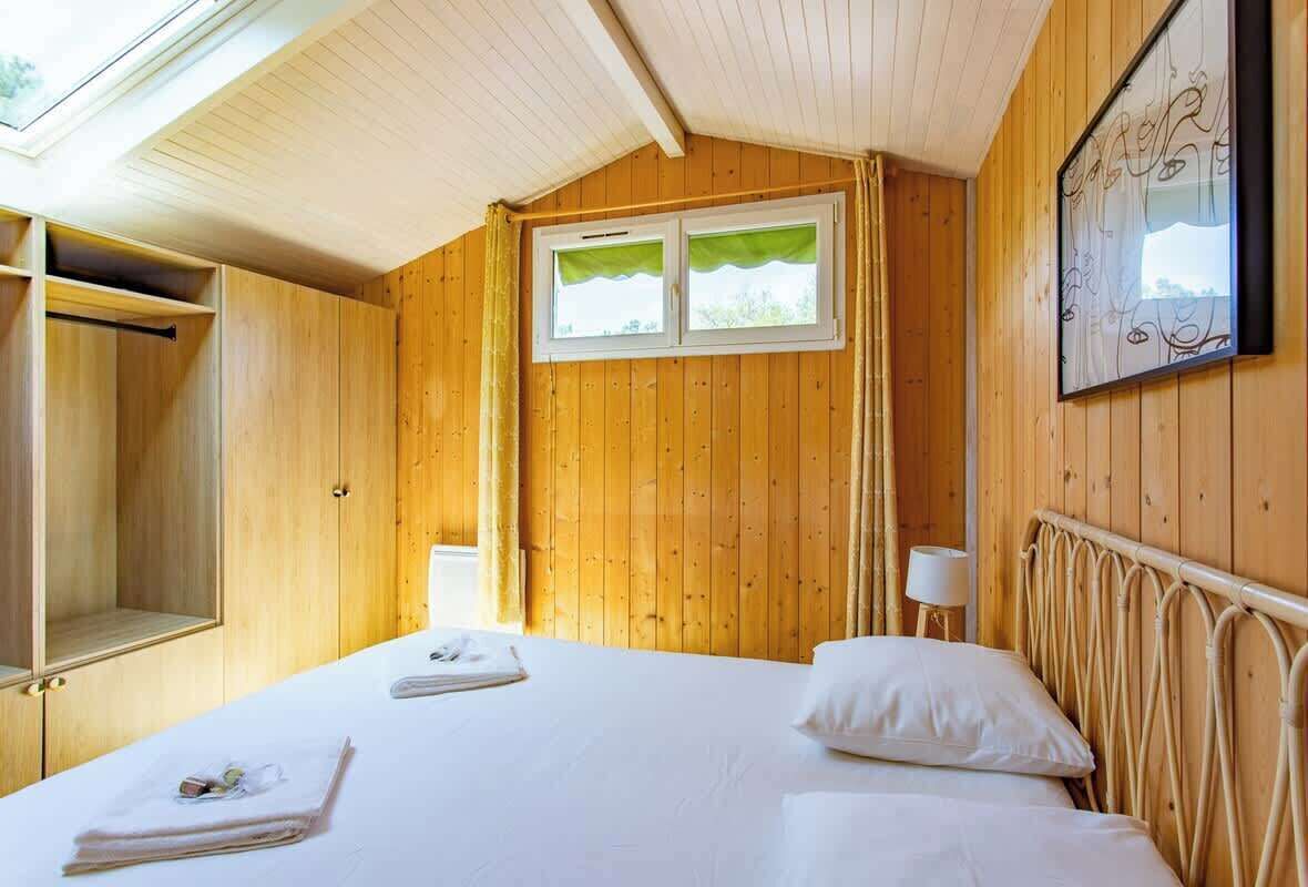 A wooden-paneled room with a bed, a window with curtains, a bedside lamp, and a wall-mounted artwork.