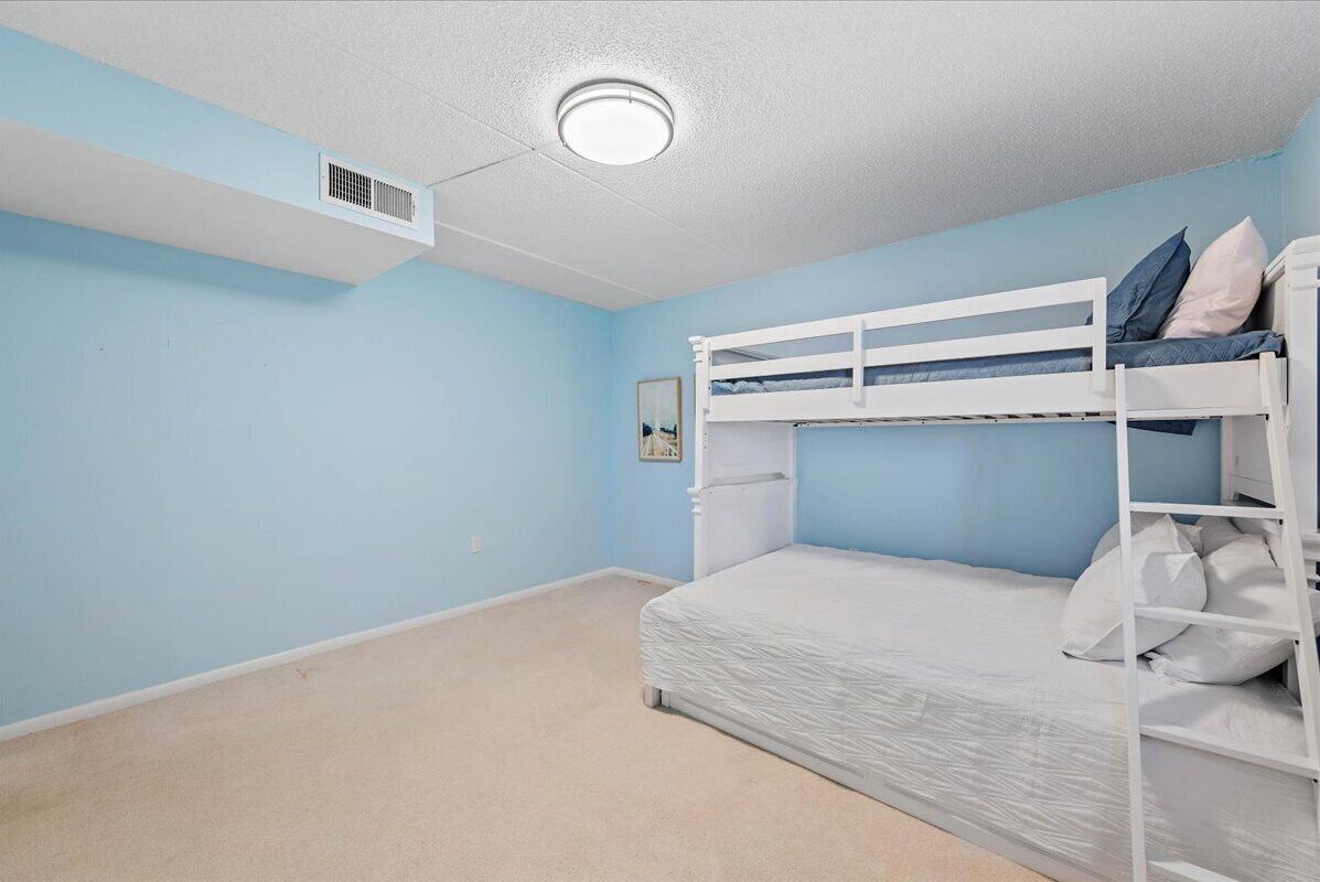 A room with a bunk bed, a single bed, a ceiling vent, and a framed picture on the wall.