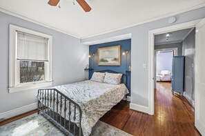 A bedroom with a bed, a window with blinds, a painting on the wall, and a door leading to another room. - Starbright Cottage by Seaside Vacations & Sales (Chincoteague)