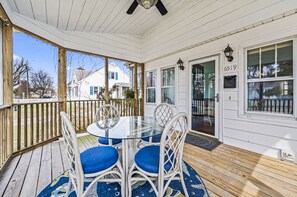 A covered porch with a glass table and wicker chairs, a wooden deck, and a white house in the background. - Starbright Cottage by Seaside Vacations & Sales (Chincoteague)