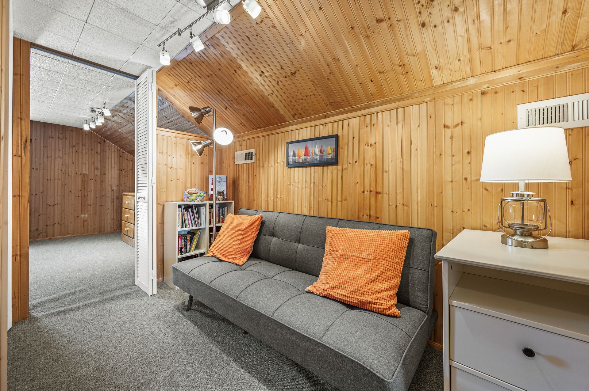 A cozy room with a grey sofa, orange cushions, a bookshelf, a desk with a lamp, and wooden walls.