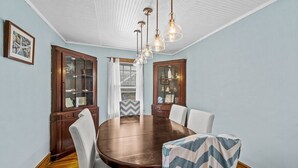 Un comedor con una mesa redonda de madera, sillas blancas y una araña. Se observan dos armarios de madera con puertas de cristal y una ventana con cortinas.