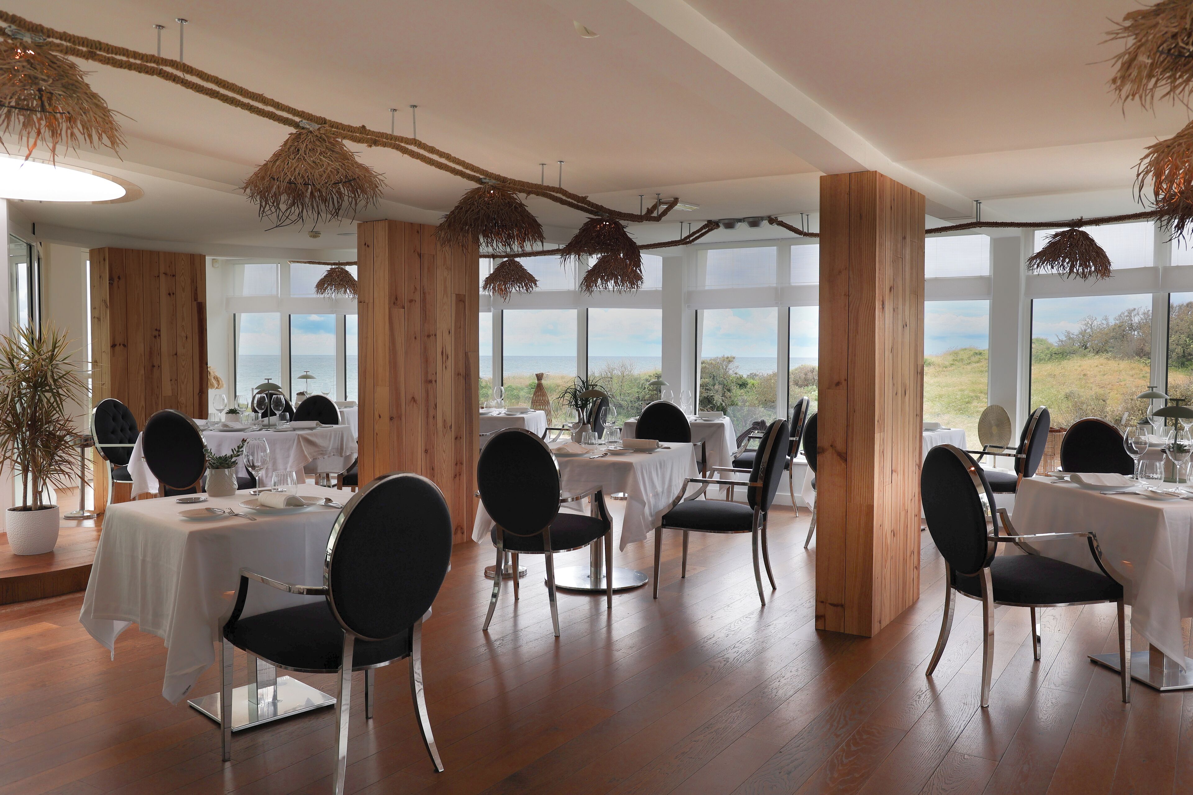 Comedor con mesas dispuestas para seis, paneles de madera y grandes ventanales con vistas al exterior.