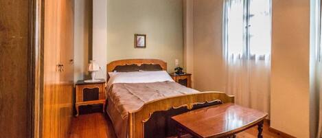 A bedroom with a wooden bed, a bedside table with a lamp, a nightstand, and a window with curtains.