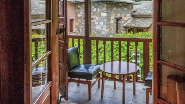 A balcony with a stone building in the background, a wooden table, two chairs, and a view of greenery. - Calliope s Stone Manor (Zagora-Mouresi)