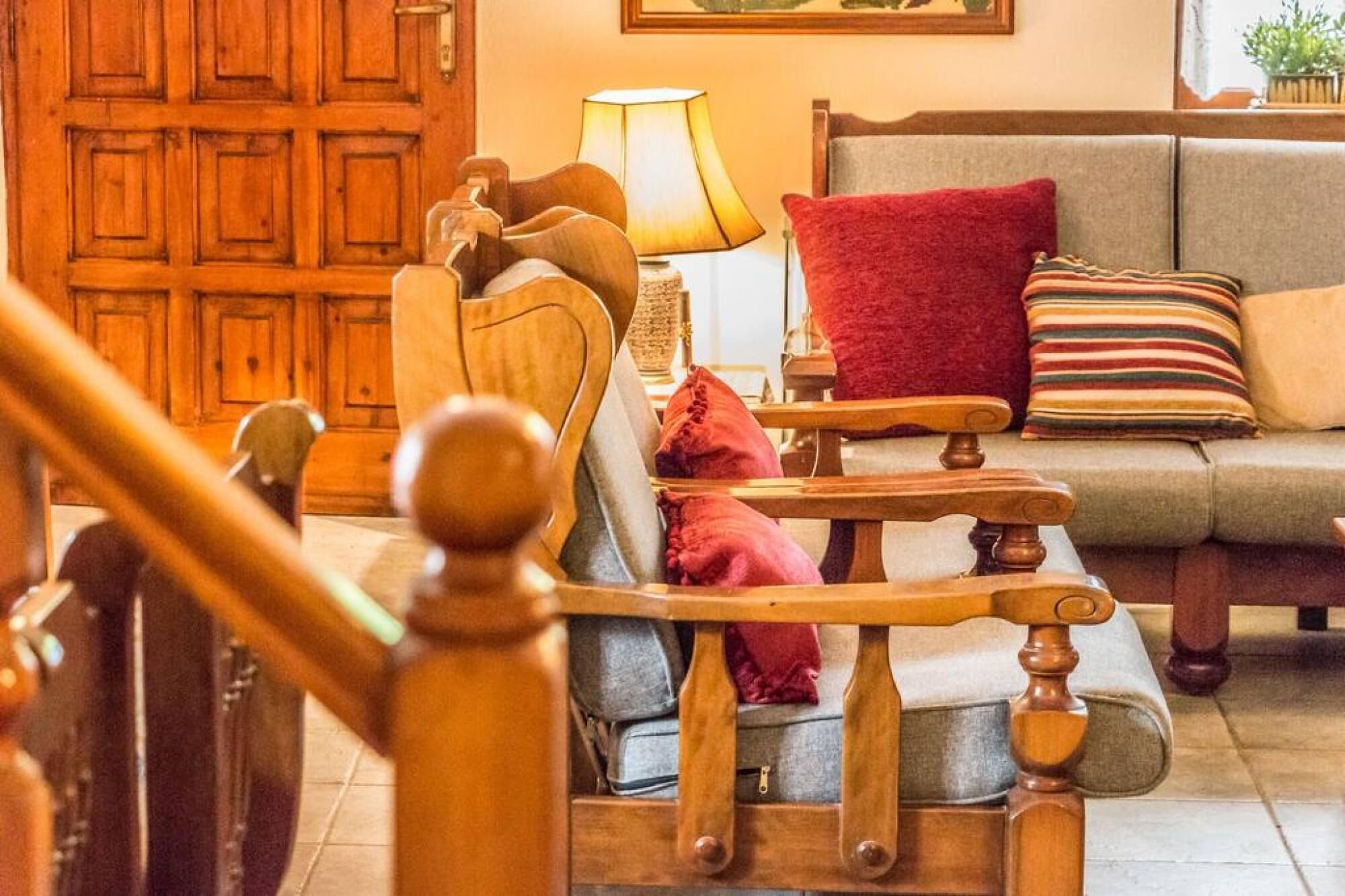 A wooden chair with cushions, a sofa with a red pillow, and a lamp in a cozy room with wooden doors.