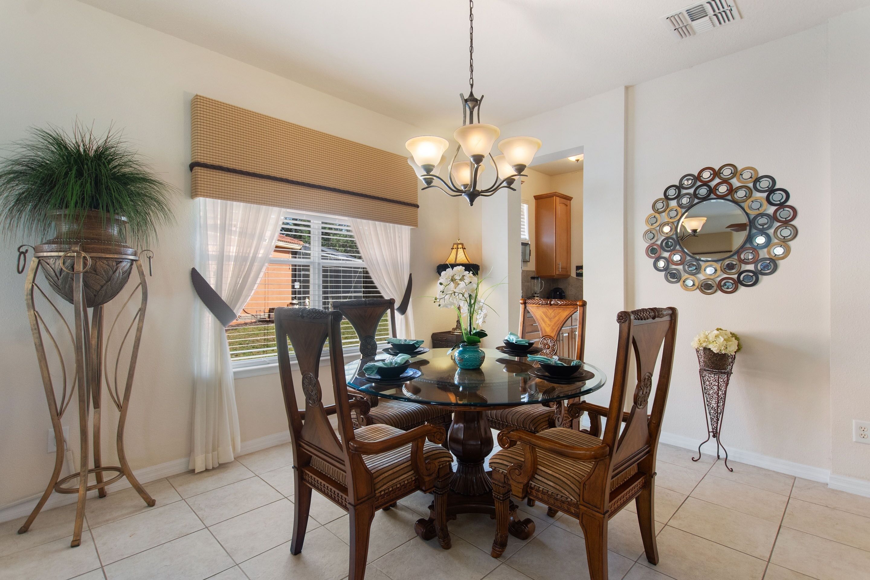 A dining area with a round glass table, six wooden chairs, a chandelier, a potted plant, and a decorative mirror on the wall.