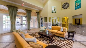 A spacious lobby with a large sofa, a coffee table, and a fireplace. There are large windows with curtains, a reception desk, and a wall with a decorative mirror and artwork.